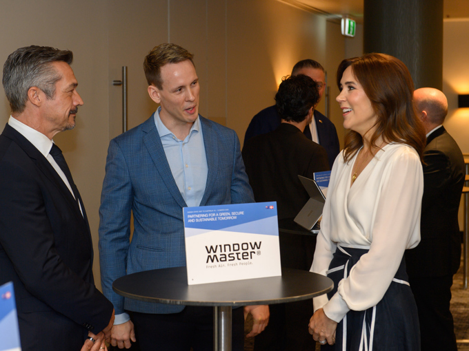 Personer i forretningsantrekk står og snakker sammen rundt et høyt bord med et «WindowMaster»-skilt på en konferanse eller et nettverksarrangement.
