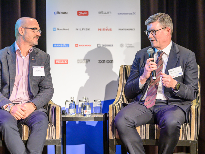 To menn i dress som diskuterer på scenen, med en mikrofon, vannflasker og et sponsorplakat i bakgrunnen.