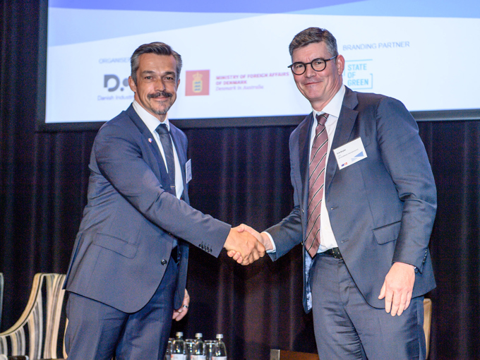 To menn i dress som håndhilser på et formelt arrangement, med en presentasjonsskjerm og vannflasker i bakgrunnen.